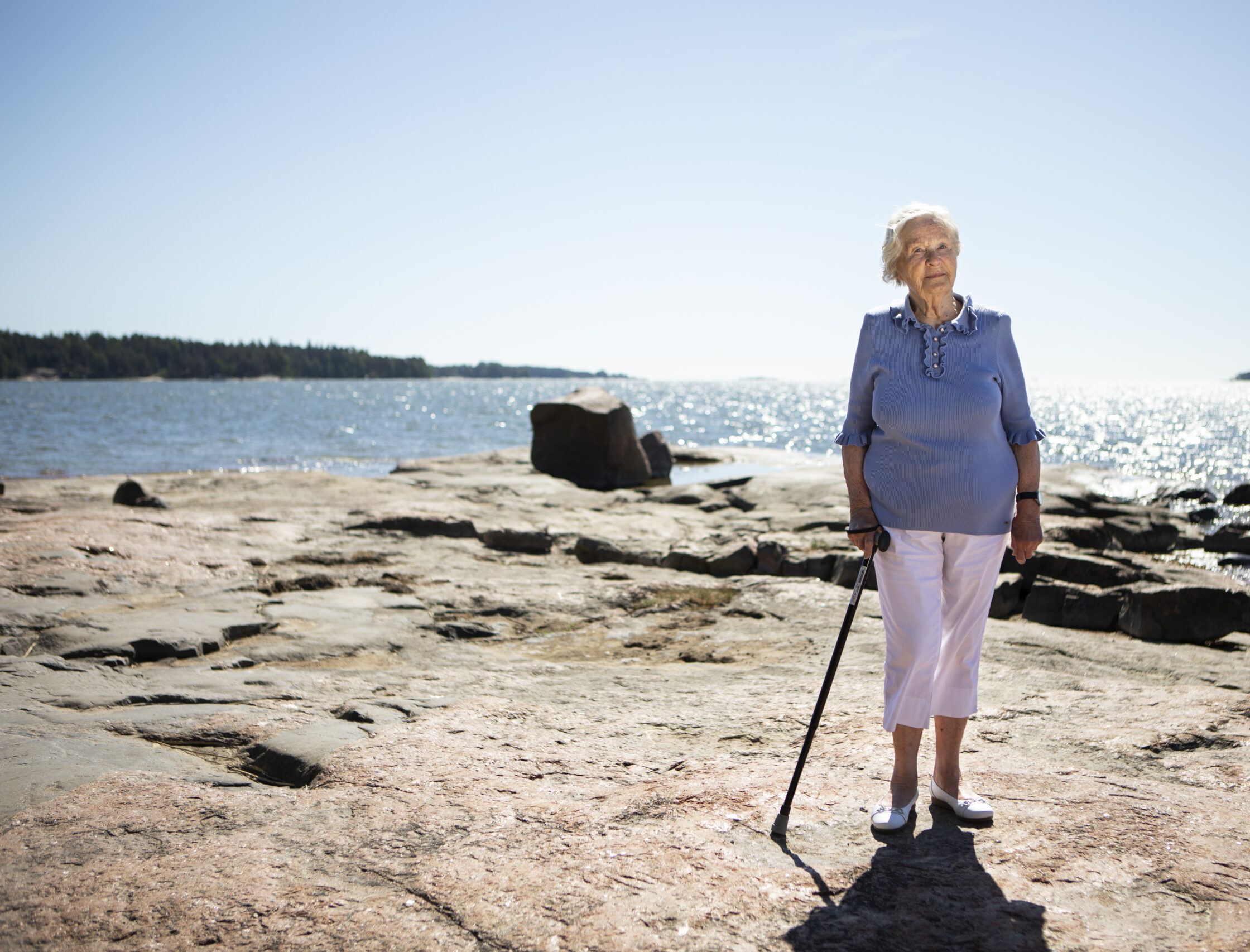 Kuvassa vaaleansiniseen puseroon ja valkoisiin housuihin pukeutunut iäkäs rouva nojaa kävelykeppiin; hän seisoo tasaisella rantakalliolla. Takana näkyy auringossa kimmeltävä meri.