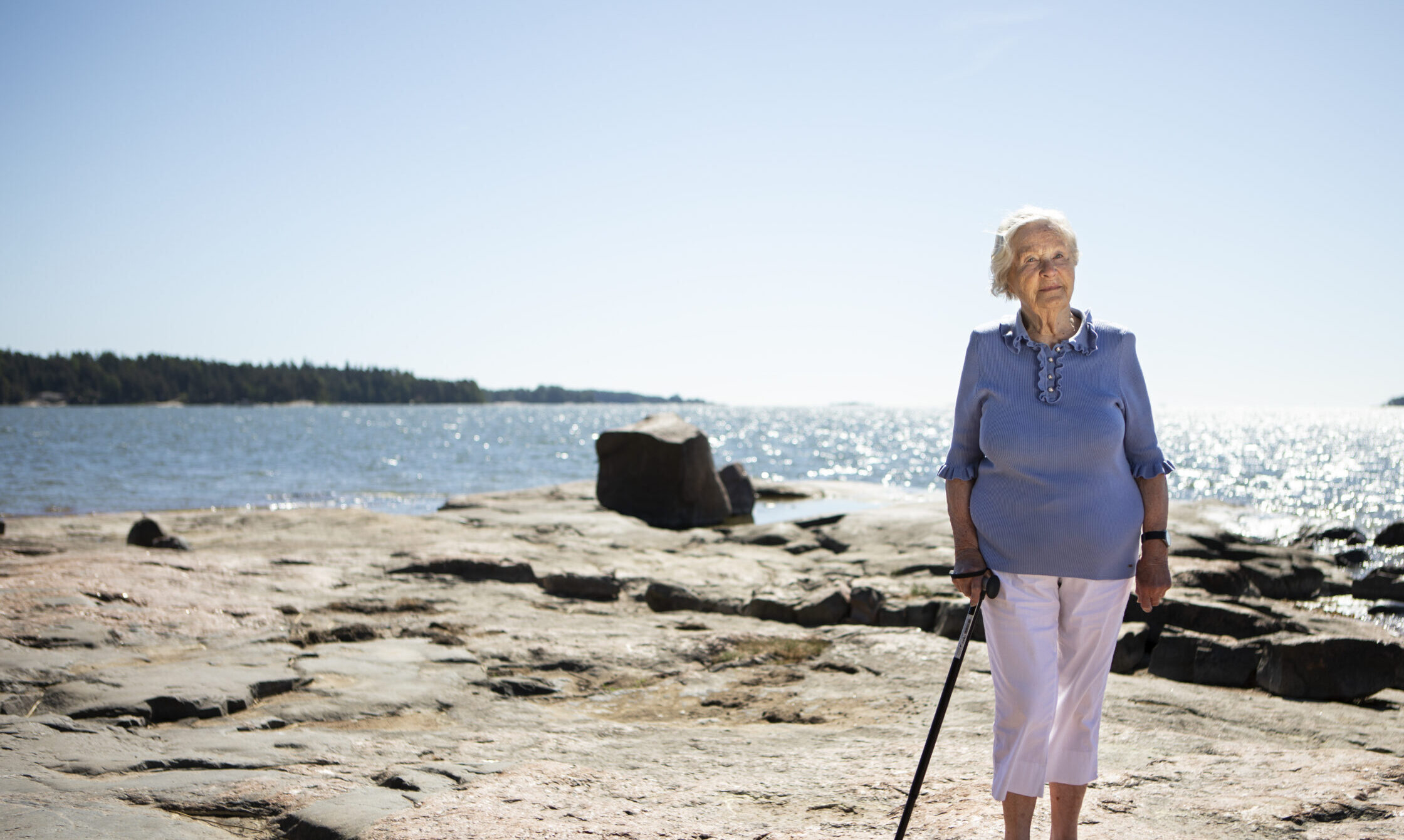 Kuvassa vaaleansiniseen puseroon ja valkoisiin housuihin pukeutunut iäkäs rouva nojaa kävelykeppiin; hän seisoo tasaisella rantakalliolla. Takana näkyy auringossa kimmeltävä meri.