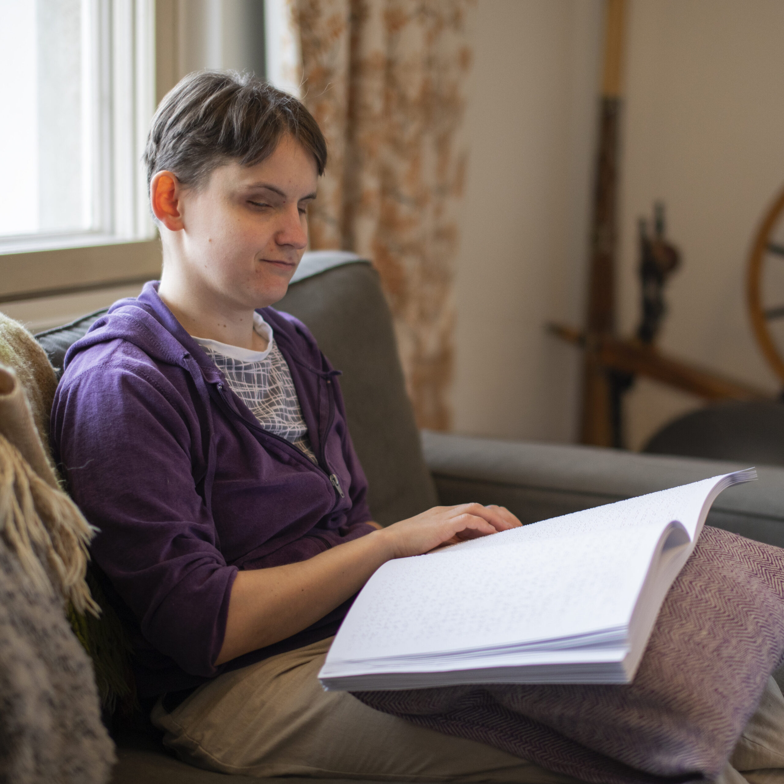 Nainen istuu sohvalla ja lukee kirjaa keskittyneesti kodinomaisessa ympäristössä, olohuoneessa sohvalla.